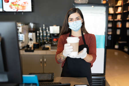Female employee wearing face mask and gloves while serving coffee in restaurantの写真素材