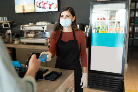 Female barista in face mask receiving payment through smartphone at coffee shopの写真素材