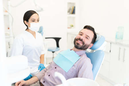 Female dentist standing by smiling mid adult man sitting on chair in clinic