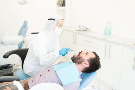 Female odontologist in protective kit examining teeth of man during coronavirus outbreakの写真素材