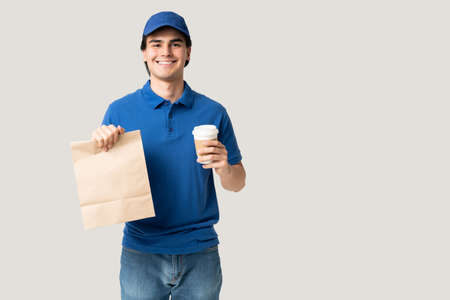 Confident handsome Latin delivery man holding disposable cup and paper bag while standing in studioの写真素材