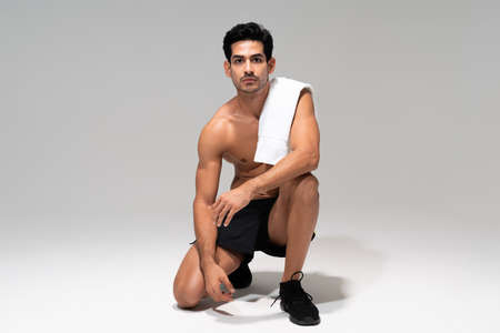Young Latin athletic man with towel on shoulder kneeling in studio with gray background