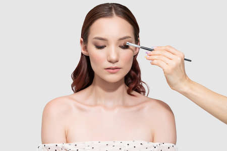 Makeup artist applying eyeshadow with brush on eyelid of beautiful young woman against gray backgroundの写真素材