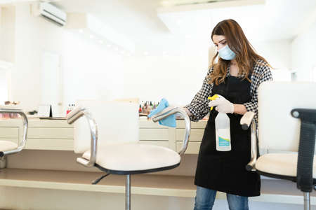 Young hairstylist wearing face mask while cleaning chair in beauty studio during coronavirus...