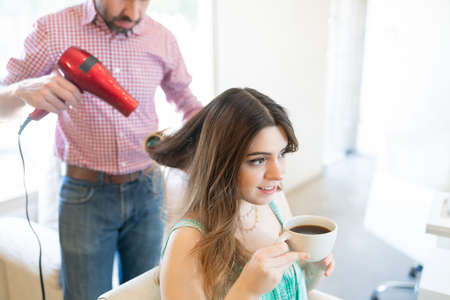 Male hairstylist using blow dryer on hair of young female customer drinking coffee at beauty salonの写真素材