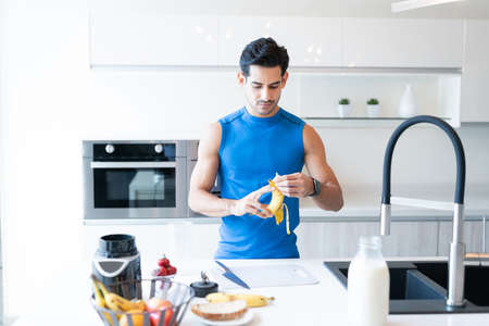 Young athletic man peeling banana while standing at kitchen islandの写真素材