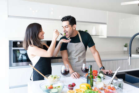 Smiling woman feeding pasta to boyfriend while standing at kitchen island