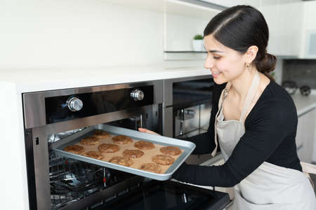 Smiling Hispanic young woman keeping tray of cookies in microwave oven