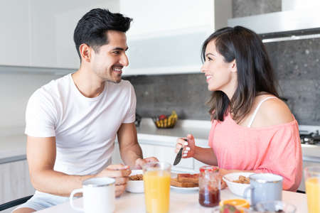 Smiling Latin young couple looking at each other while having breakfast in kitchenの写真素材