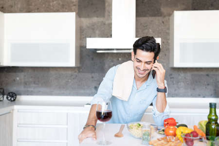 Smiling young man talking on mobile phone while preparing food at homeの写真素材