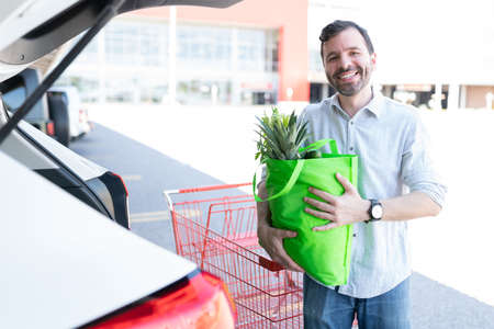 Smiling Hispanic man carrying grocery bag while standing by car parked outside supermarketの写真素材