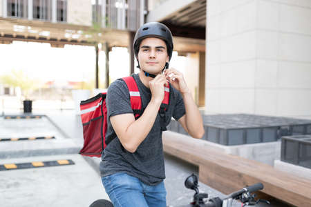 Confident food delivery man adjusting belt of helmet while sitting on bicycleの写真素材