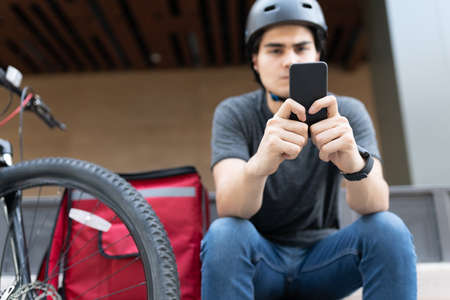Young delivery man using app on smartphone while sitting by bicycleの写真素材