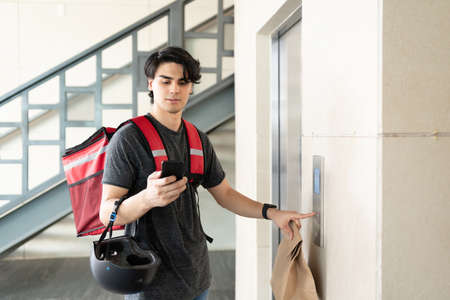 Latin delivery man using smartphone while pressing elevator button in building