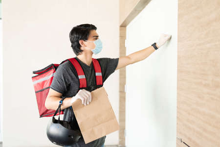 Young delivery man in face mask knocking door while holding parcel at apartmentの写真素材
