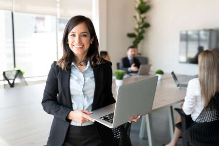 Portrait of smiling female entrepreneur holding a laptop with team in background at office conference roomの写真素材