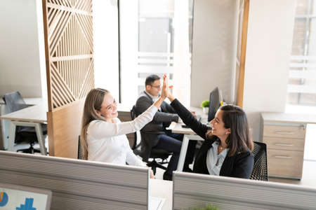 Two successful female colleagues giving high-five to each other while sitting at desk in workplace