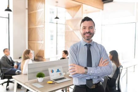 Portrait of a white collar worker standing in front of his desk at office with colleagues working in backgroundの写真素材