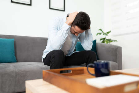 Latin young man having a headache while sitting on a sofa after a busy day at work