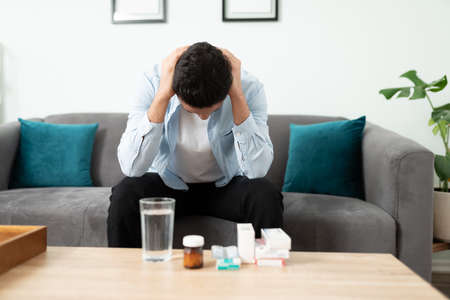 Man in his 30s sitting on a sofa at home with a too many pills to take and feeling overwhelmed and...