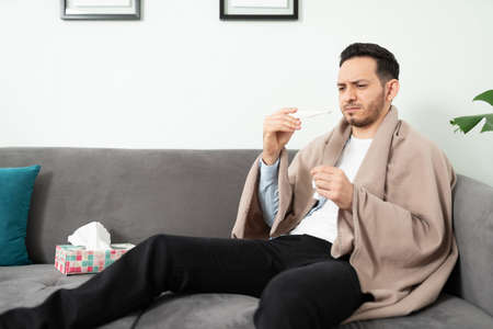 Good looking man feeling sick while resting on a sofa while looking at a thermometer