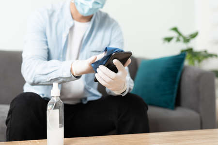 Closeup of a man wearing a face mask and disinfecting his phone while staying home during...
