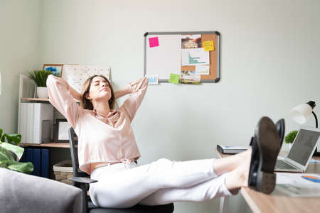 Full length view of a woman dressed casual and relaxing in her office with her feet up the desk and leaning back on her chairの写真素材