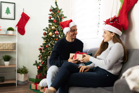 Attractive Latin man handing over a Christmas gift to his partner while celebrating together at home