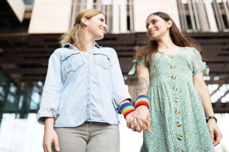 Low angle of a caucasian gay woman and a latin lesbian woman standing and holding hands with rainbow colored wristbandsの写真素材