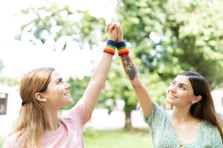 A pair of attractive young women friends with their hands in the air and wearing LGBT rainbow colored wristbandsの写真素材