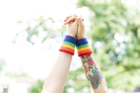 Close up of two arms raised holding hands in the air and wearing rainbow wristbands in support of...