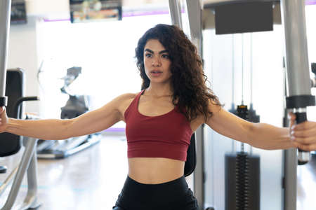 Beautiful young woman with curly hair working out in a chest machin at the gym