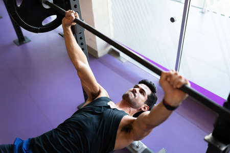 Fit young man seen from above fixed on lifting a heavy barbell and lying on a gym bench