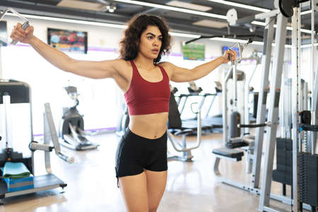 Latin young woman stretching her arms in a cable machine to exercise her chest muscles in the gym