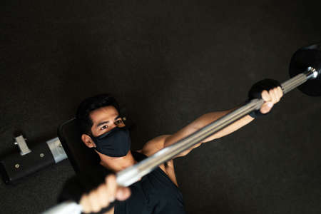 High angle of a hispanic young man using a face mask training with weights in a barbell at the fitness club during the pandemicの写真素材
