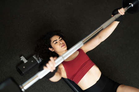 Hispanic young woman seen from above lying on a bench and doing a weight lifting workout with a barbell at the gymの写真素材