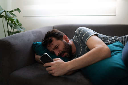 Brokenhearted man in his 30s lying on the couch looking depressed and waiting for a call or message on his cellphoneの写真素材