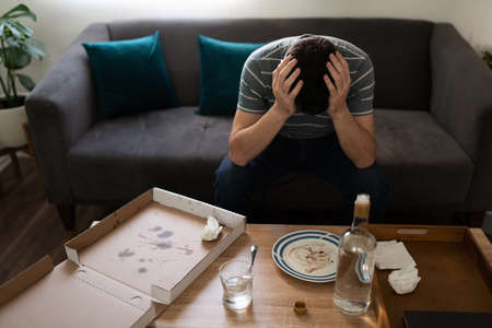 Sad adult man sitting on the living room and holding his head with the hands in front of a table...