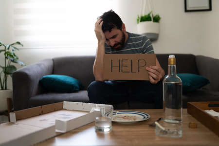Depressed and alcoholic latin adult man sitting alone on the sofa and holding a sign with help...