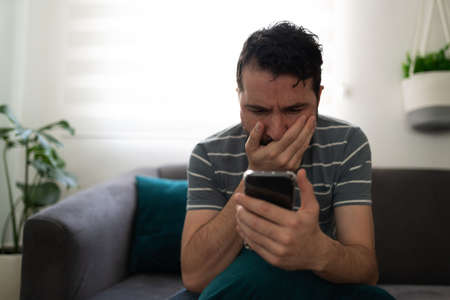 Latin man looking surprised while looking at his cellphone and sitting alone on the couch