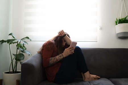 Latin young woman suffering from loneliness sitting on the couch with her cellphone. Woman in her...