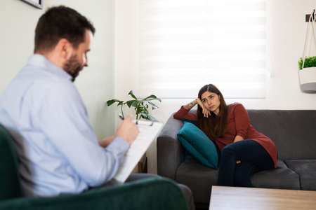 Young latin woman getting professional psychological therapy for her depression and anxiety. Male psychologist writing in his chart during a session with a female patientの写真素材