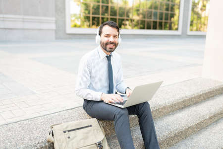 Hispanic professional man with headphones working on his computer outside a city buildingの写真素材