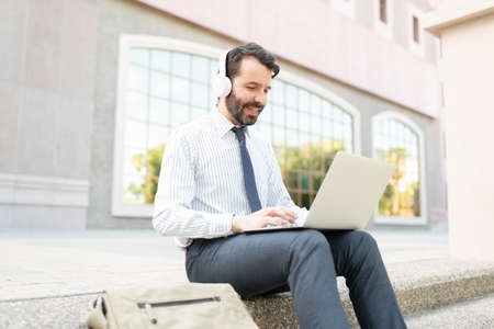 Side view of an attractive male worker happily listening to music and using a laptop outside an office buildingの写真素材