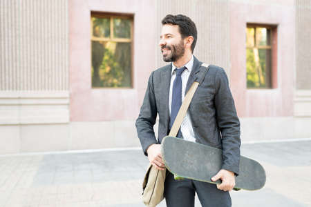 Attractive man in his 30s smiling and holding his skateboard outside a building. Latin worker using...