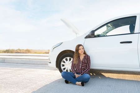 Sad young woman sitting on the road next to his broken car with the hood open and waiting for road assistance helpの写真素材