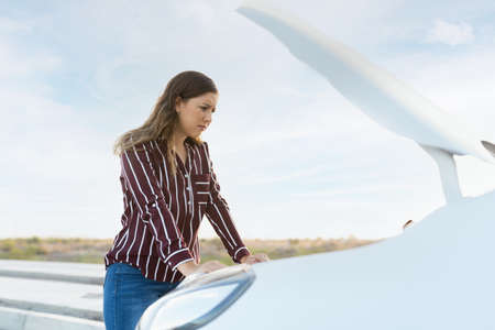 Caucasian young woman with her car hood open and looking at the broken-down engine with a sad and...