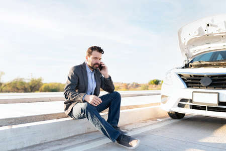 Latin man sitting on the roadside and talking on the phone to ask for help and a tow truck. Man in...