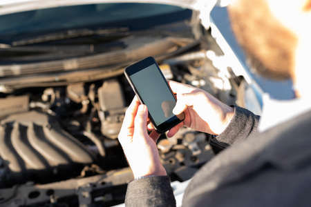 Adult man in his 30s with his car hood open and texting a mechanic because his car broke down in the middle of the highwayの写真素材