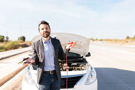 Hispanic man in his 30s holding the jumper cables looking upset because his car battery is flatの写真素材